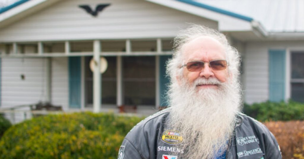 An older white man with a white beard stands in front of a white home