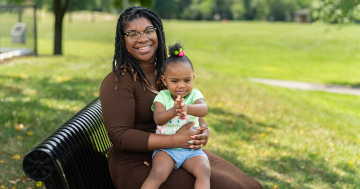 A smiling, young black woman sits on a bench holding a black toddler.