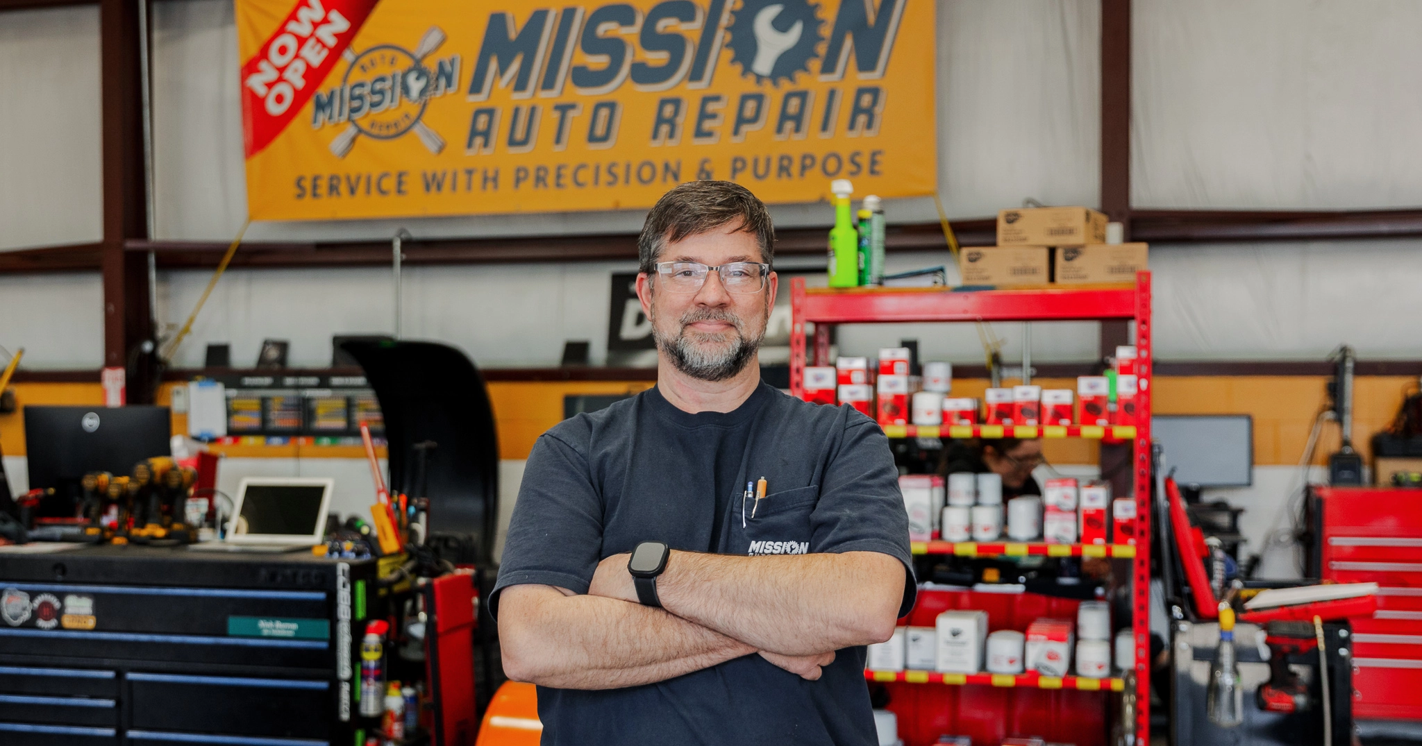 A white man with folded arms stands in a auto mechanic garage in front of tools