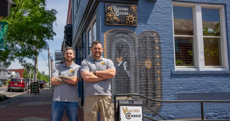 Two men in grey polo shirts stand outside a blue painted brick building with a sign that says "Vaulted Cards"