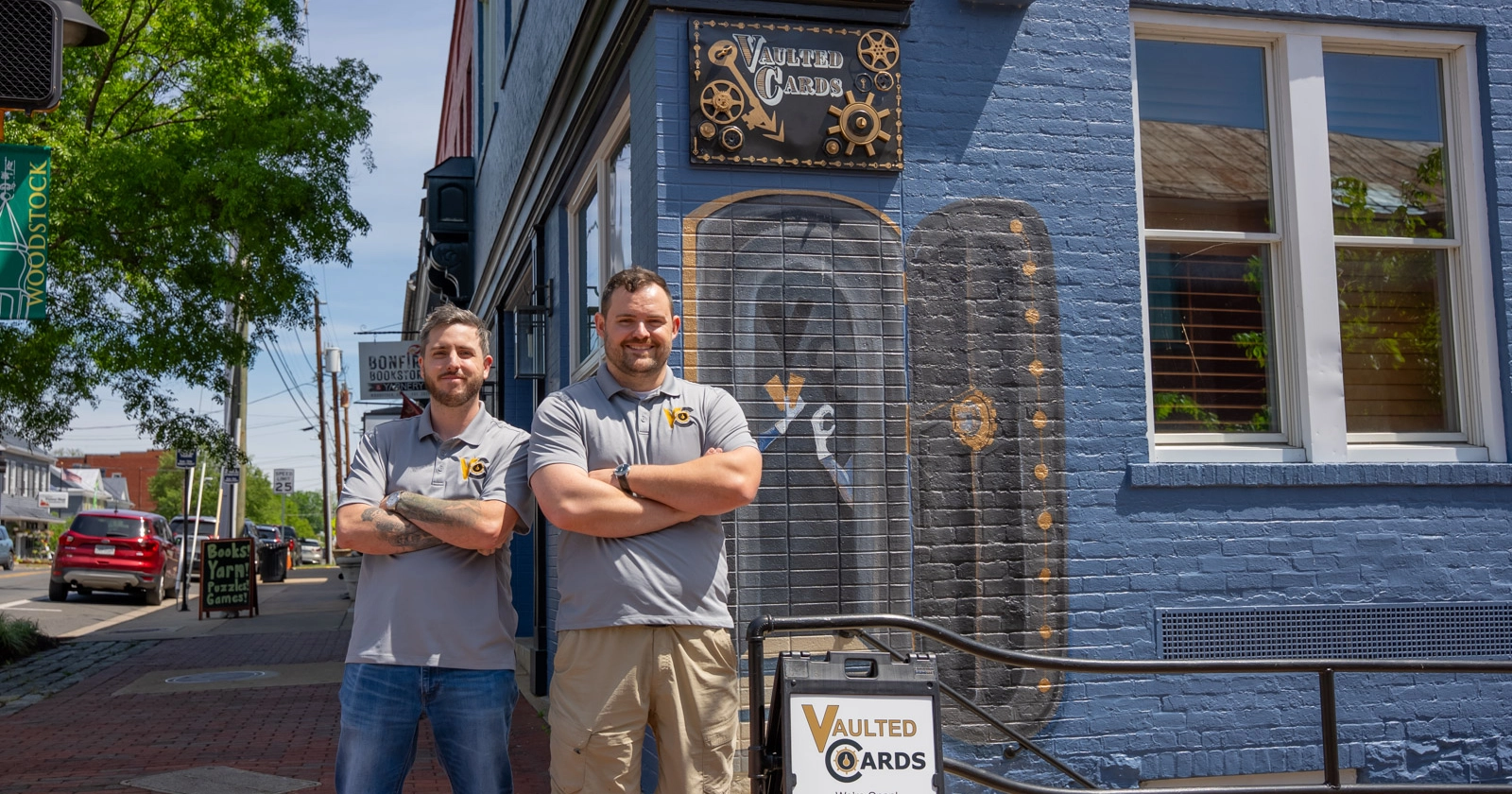 Two men in grey polo shirts stand outside a blue painted brick building with a sign that says "Vaulted Cards"