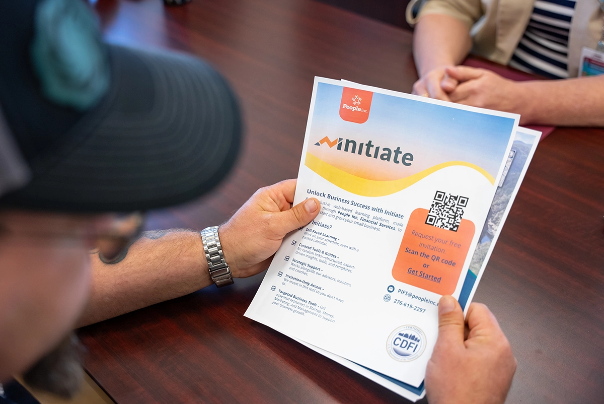 A man wearing a black baseball hat holds a flyer for the Initiate program with People Incorporated Financial Services.