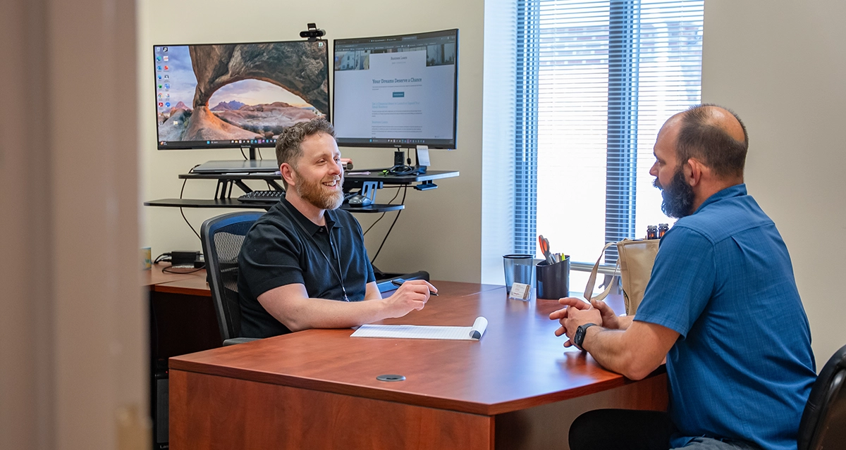 Two white men sit across from each other in the People Inc Financial Services Office. The man on the left sits behind a large brown desk with a monitor behind him. He is wearing a black shirt and smiling. He is taking notes The man on the right is wearing a blue shirt.